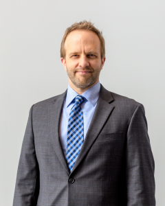 Scott Cryer wearing suit and blue tie smiling at camera with grey background