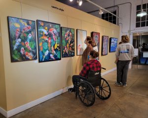 Two patrons, one standing and one in a wheelchair, look at three colorful paintings of koi fish in a pond full of bright green lily pads hanging on a yellow gallery wall.