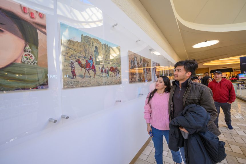 A woman in a pink sweater and a man in a black jacket look at a painting of people riding camels in front of a building