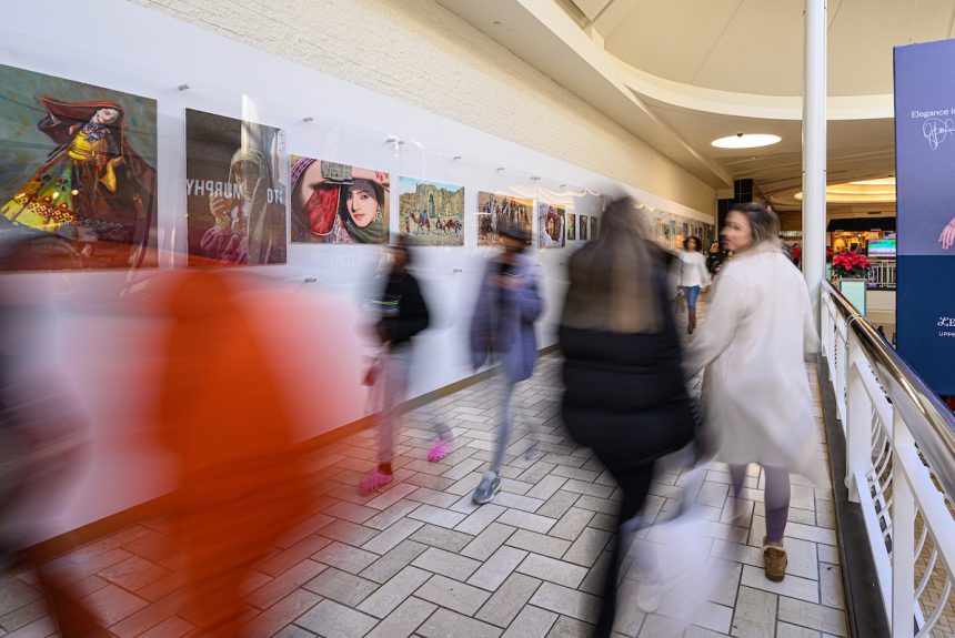In the foreground shoppers are blurry as they walk past a gallery wall of more than a dozen images in a row behind plexiglass on the wall.