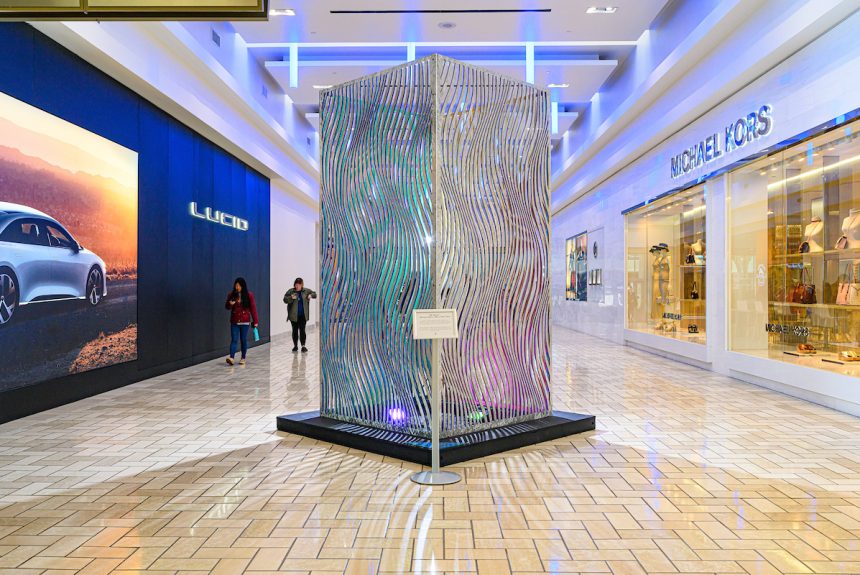 5′ wide x 10′ tall metal box with undulating, wave-like slits through which colors shimmer is standing in a mall hallway with stores on either side