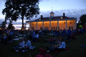 It is dusk. People sit on blankets on a lawn in front of Mount Vernon, which has its porch lit against a sunset sky