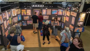 The photo looks down at a vendor display with three walls is covered with colorful canvases as patrons look at the art