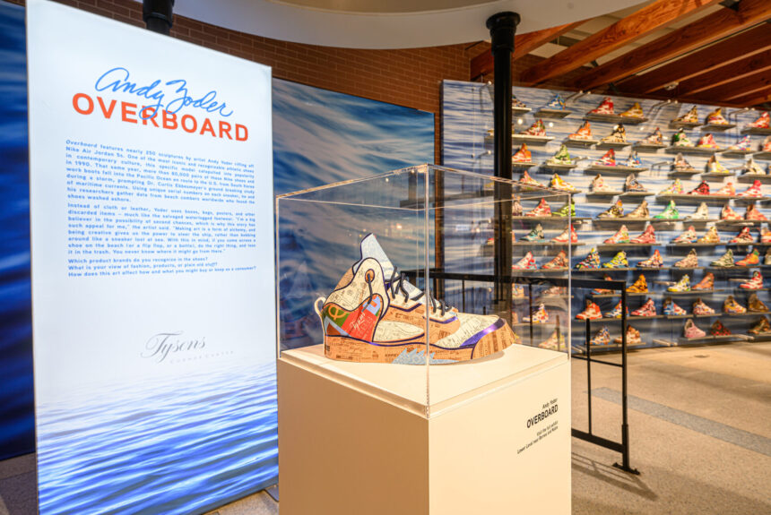In the foreground a sneaker made of recycled paper products is encased on a pedestal; in the background is an exhibition sign and a wall display of more sneakers