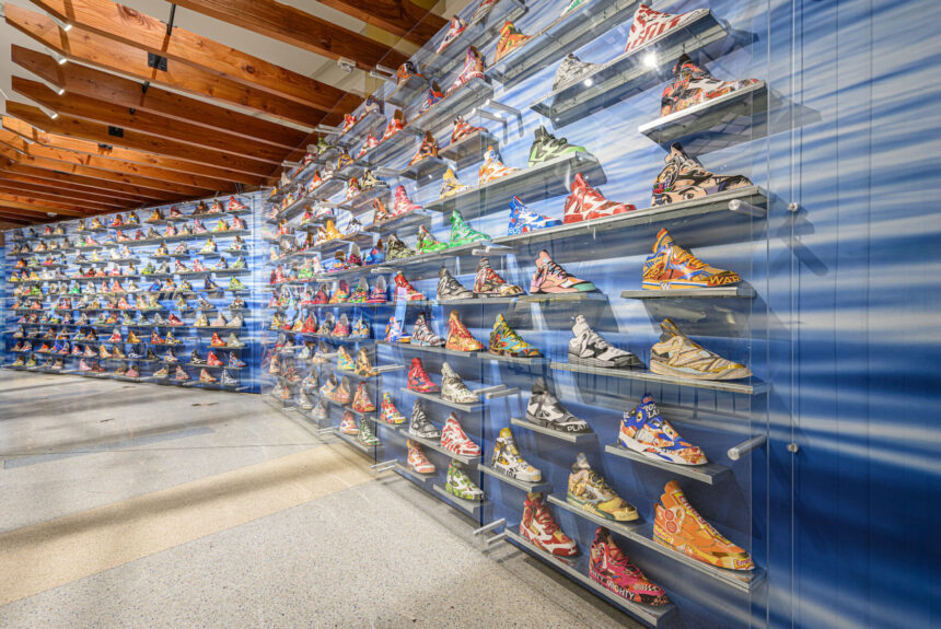Behind plexiglass on a wall with the image of rippled water are several metal shelves that each hold a model Air Jordan 5 made of recycled materials