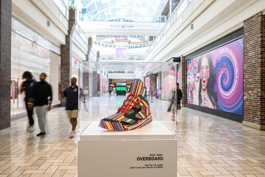 A model Air Jordan 5 made of recycled materials featuring multicolored bright stripes sits in a plexiglass case on a pedestal in the middle of a shopping center corridor.