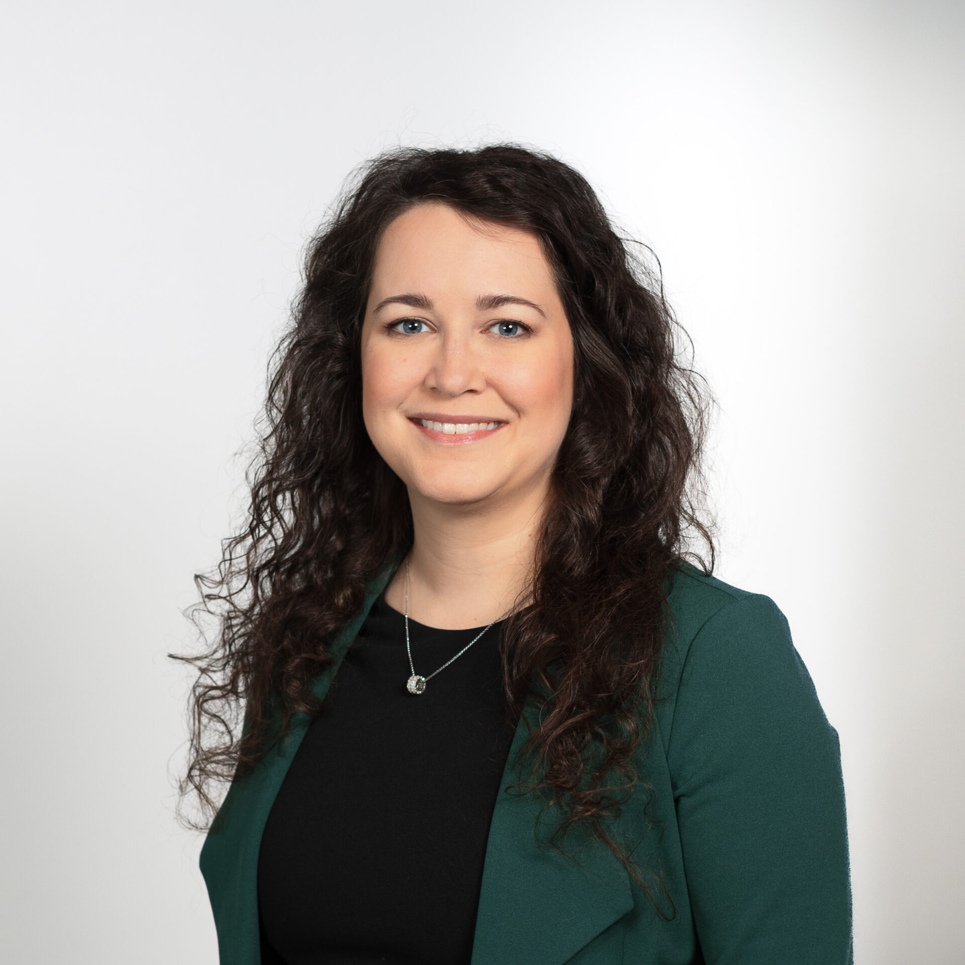 A smiling woman with long, curly dark hair wearing a black shirt, a dark green suit jacket, and a necklace