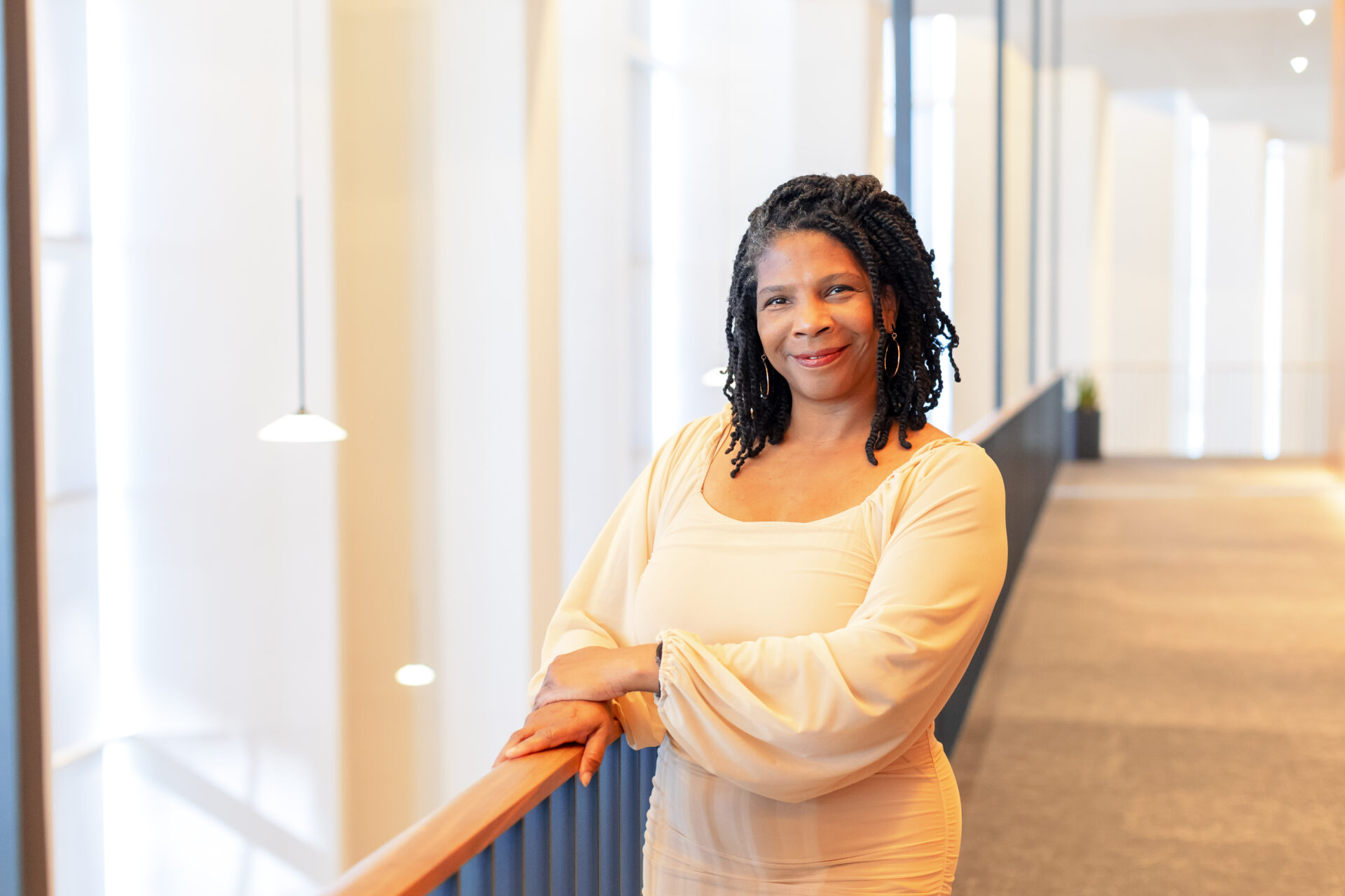 a black woman with black shoulder length braids and red lipstick wears an off-white long-sleeved dress with her hands resting on a handrail along the right side of her body.