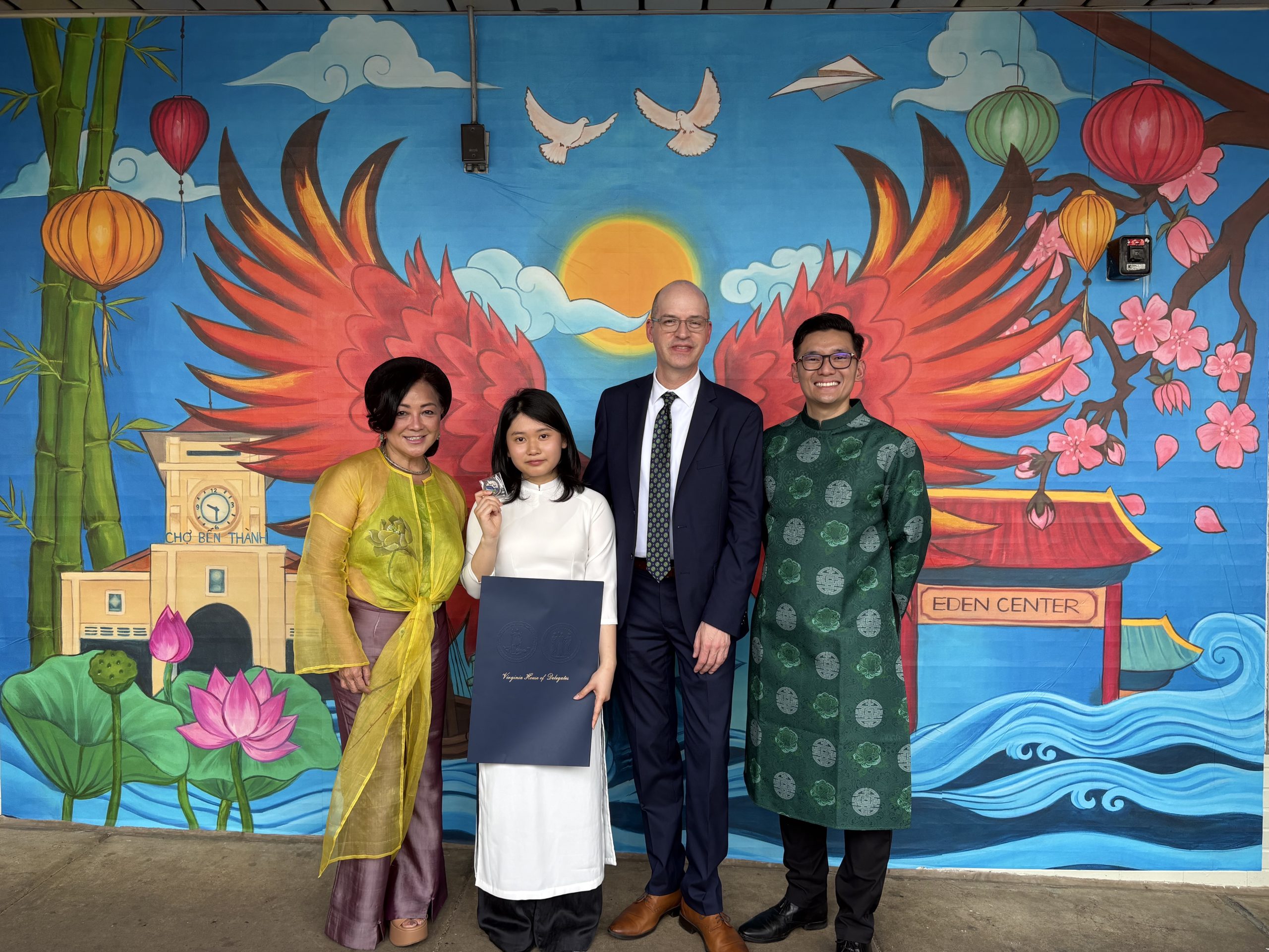 Four people pose in front of a large mural that features red phoenix wings, a rising sun, a lotus flower, and images of images of Chợ Bến Thành market in Ho Chi Minh City, Vietnam, and Eden Center at the crossroads of Seven Corners.