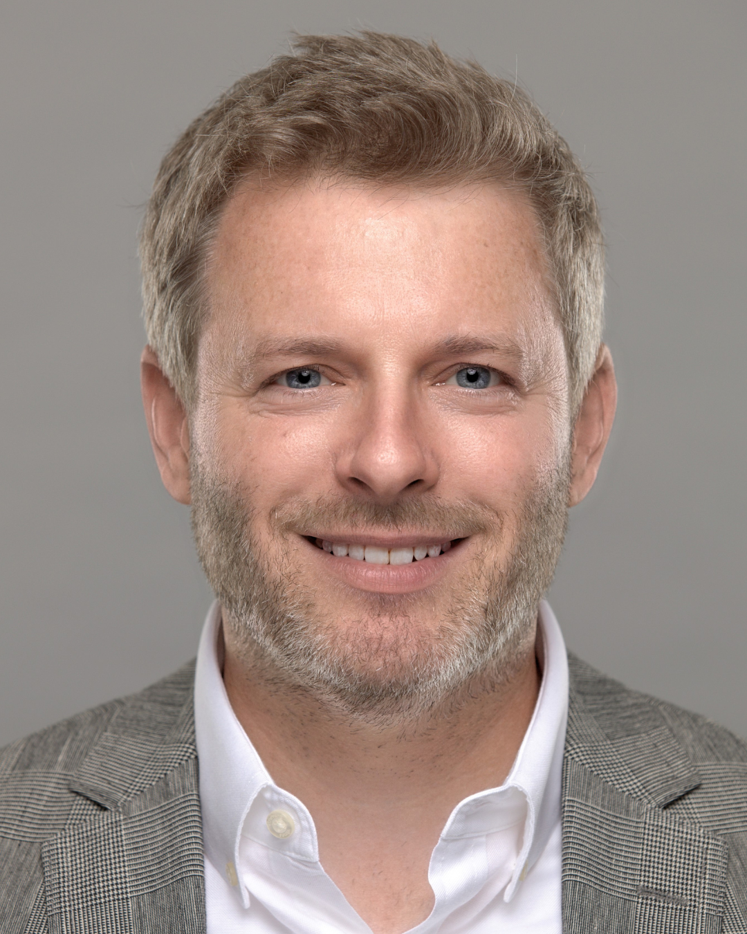 A man smiles in a headshot photo. He is wearing a white button shirt with a gray suit jacket.