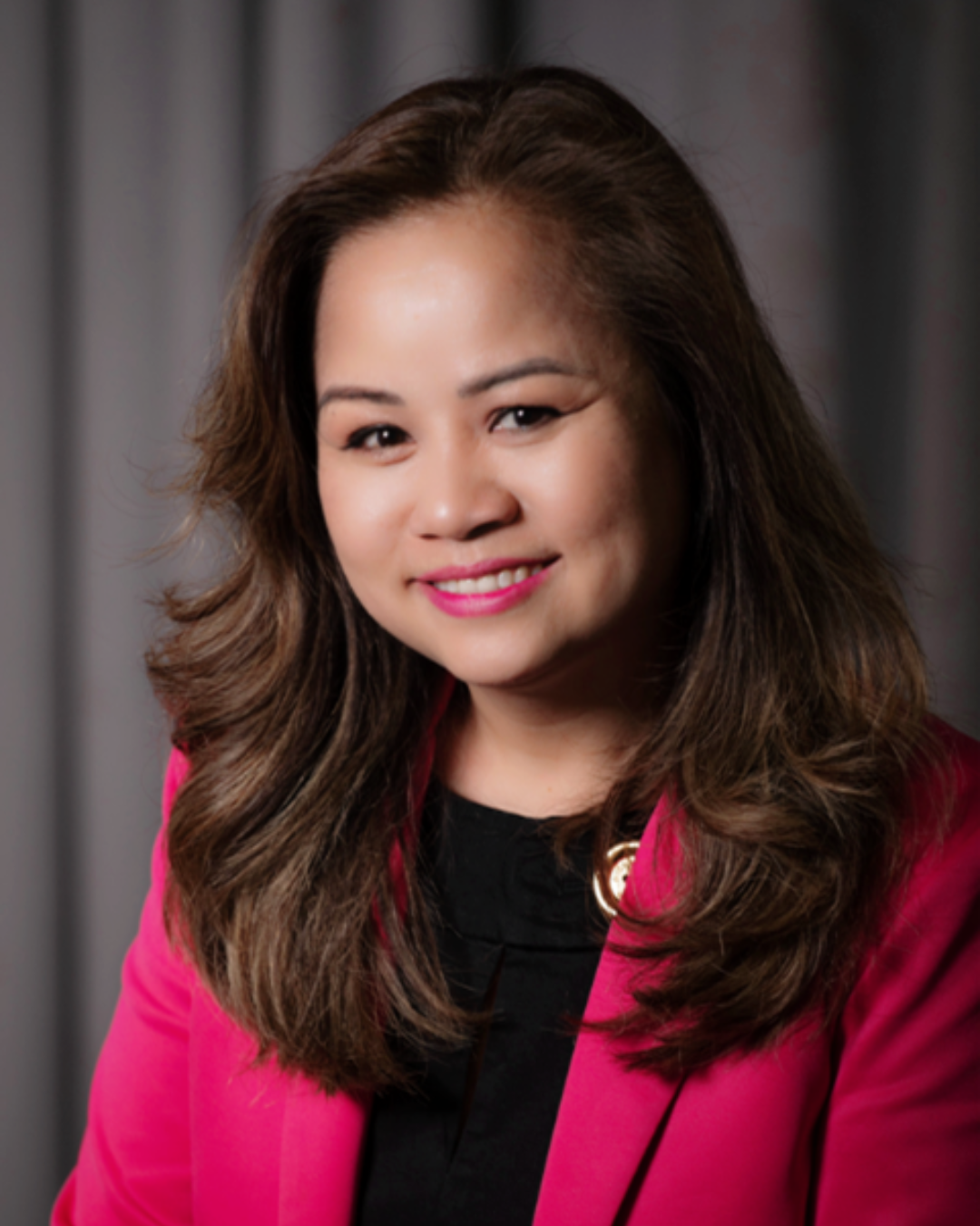 A woman smiles in a headshot photo. She has medium length dark brown hair and is wearing a black shirt with a dark pink blazer jacket.