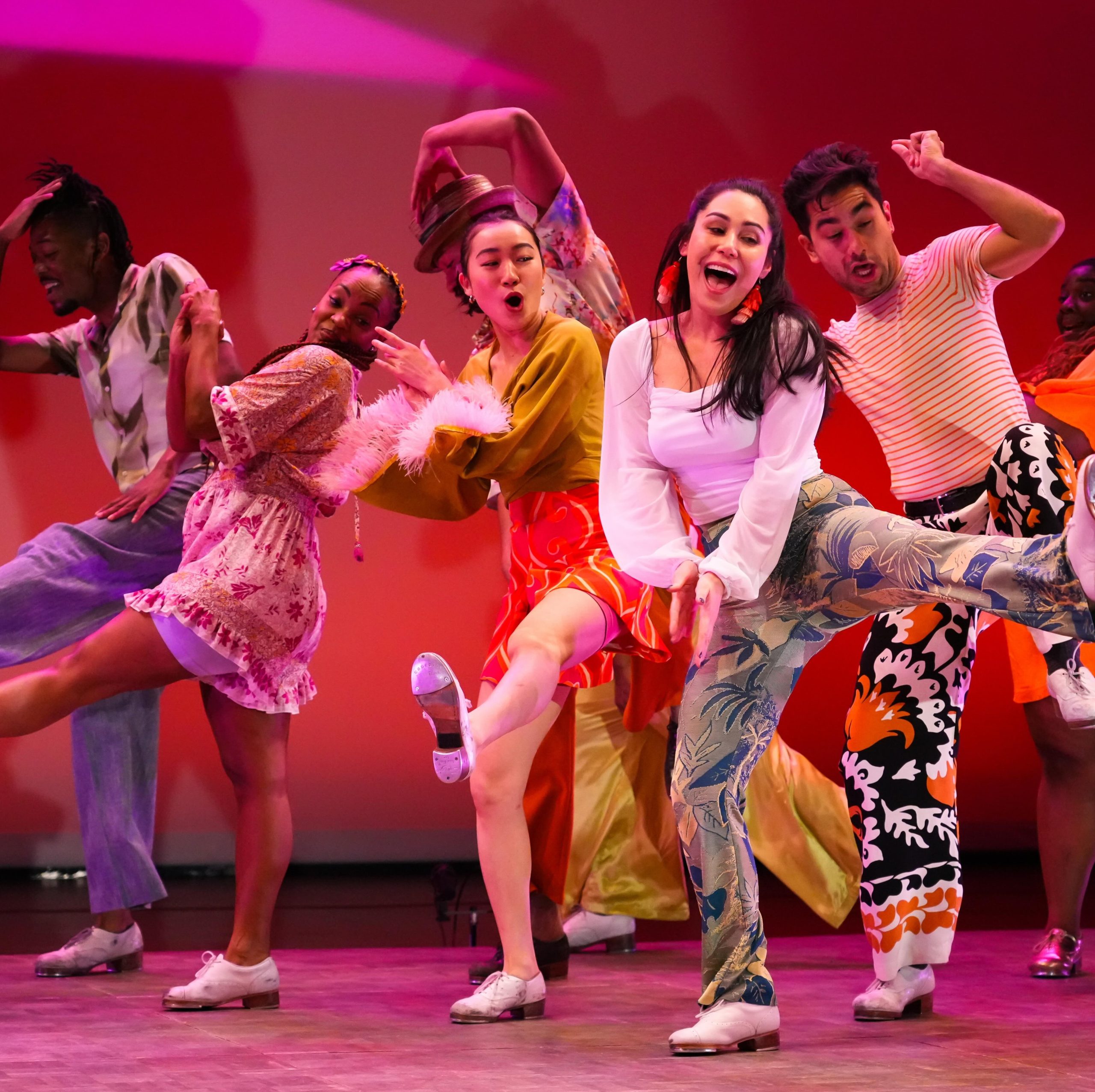 Men and women dancers in colorful clothings are dancing happily smiling with a pink and red backdrop on the stage.