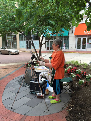 A woman wearing an orange shirt and jeans paints on a canvas surrounded by tress in Falls Church.