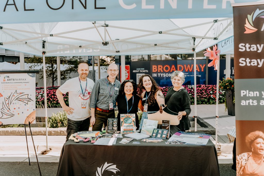Five people stand smiling outside under a vender booth tent behind a table covered in marketing materials.