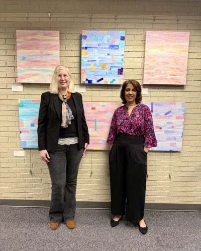 Artist Catalina Novac and Braddock Supervisor Rachna Sizemore Heizer smile in front of Novac's exhibit "Art of Happiness" featuring colorful abstract paintings at the Braddock District Supervisor's Office.